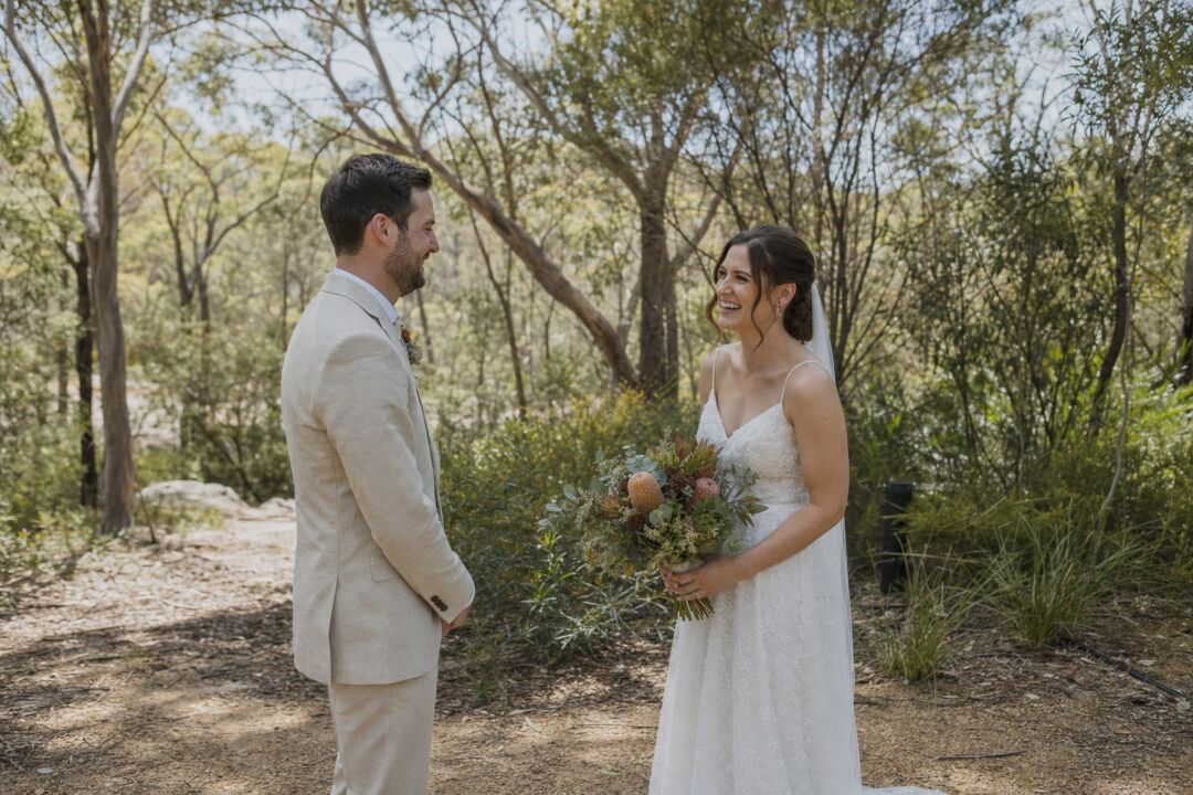A bride in a white dress and groom in a cream linen suit stand facing each other in a wooded setting.