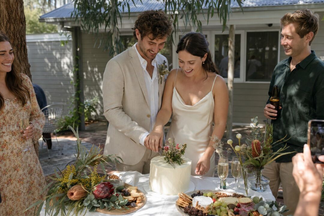 A bride and groom cut a simple white cake at their outdoor wedding reception under a willow tree.