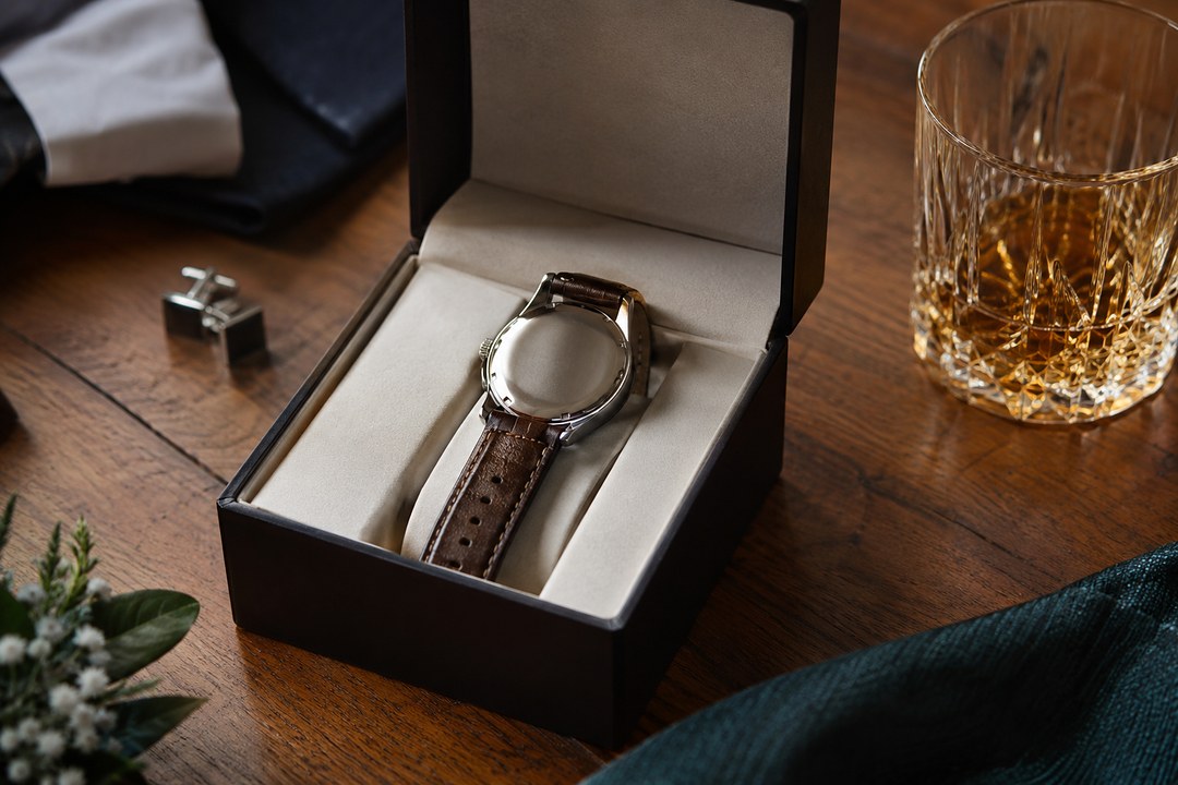A wristwatch with a brown leather strap rests face down in an open black gift box beside a whiskey tumbler and silver cufflinks.