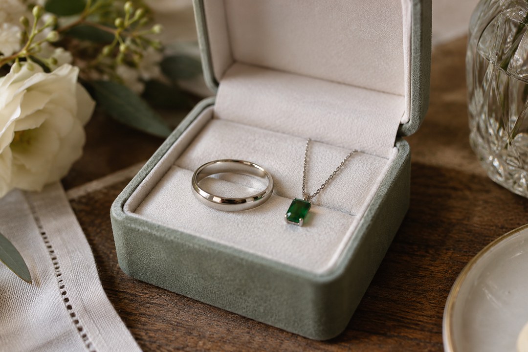A platinum band and emerald pendant rest in a sage green velvet box with white flowers and crystal on a dark wooden table.