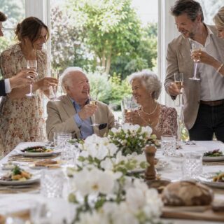 Elderly couple toast with sparkling wine at a white linen table with orchids while four adults stand in a sunlit garden room.