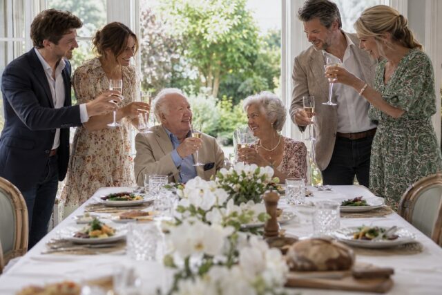 Elderly couple toast with sparkling wine at a white linen table with orchids while four adults stand in a sunlit garden room.