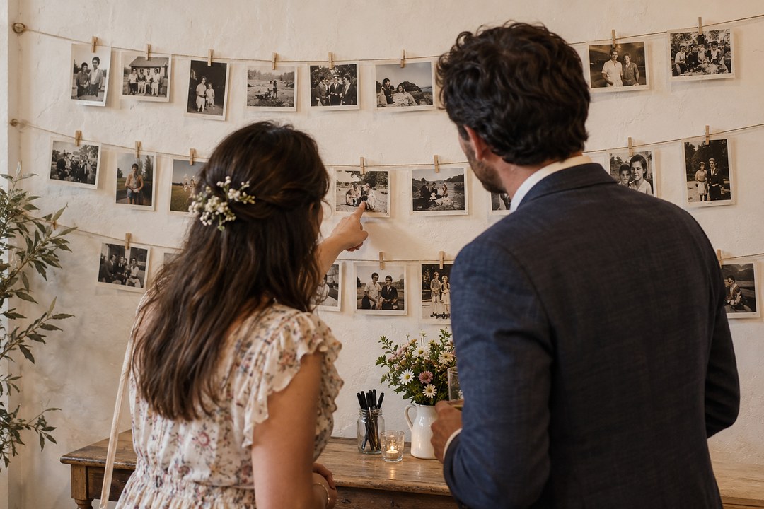 A woman in a floral dress and a man in a navy jacket look at snapshots pegged to three lines of twine on a white wall.
