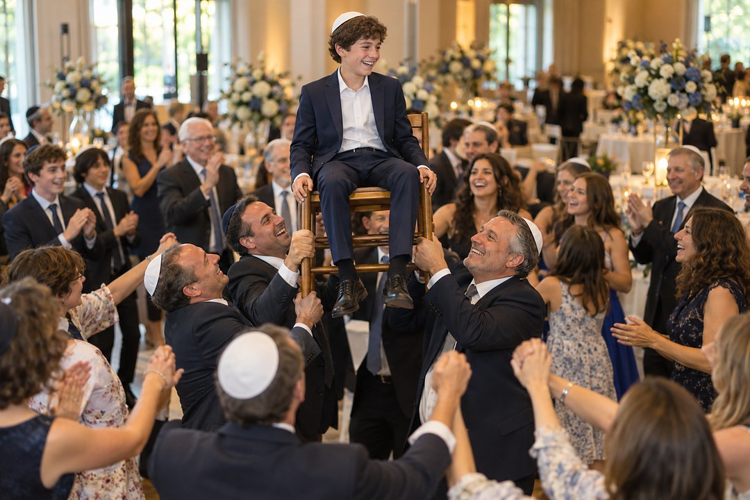 A smiling boy in a white kippah sits on a wooden chair lifted by men in dark suits above a dance floor with blue and white flowers.