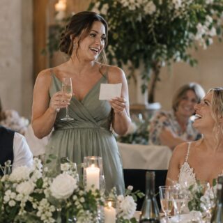 A bridesmaid in sage green holds wine and a cream card while speaking as the bride laughs near white flowers and candles.