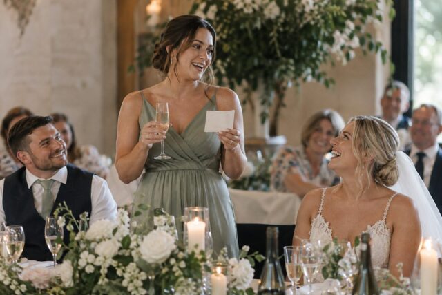 A bridesmaid in sage green holds wine and a cream card while speaking as the bride laughs near white flowers and candles.