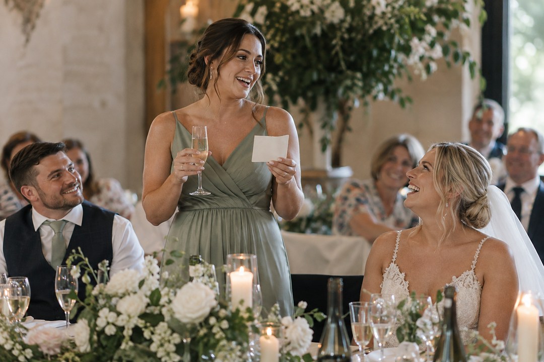 A bridesmaid in sage green holds wine and a cream card while speaking as the bride laughs near white flowers and candles.