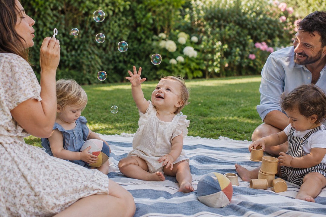 A baby in a cream romper reaches toward soap bubbles while sitting on a blue and white striped blanket as a woman blows more.