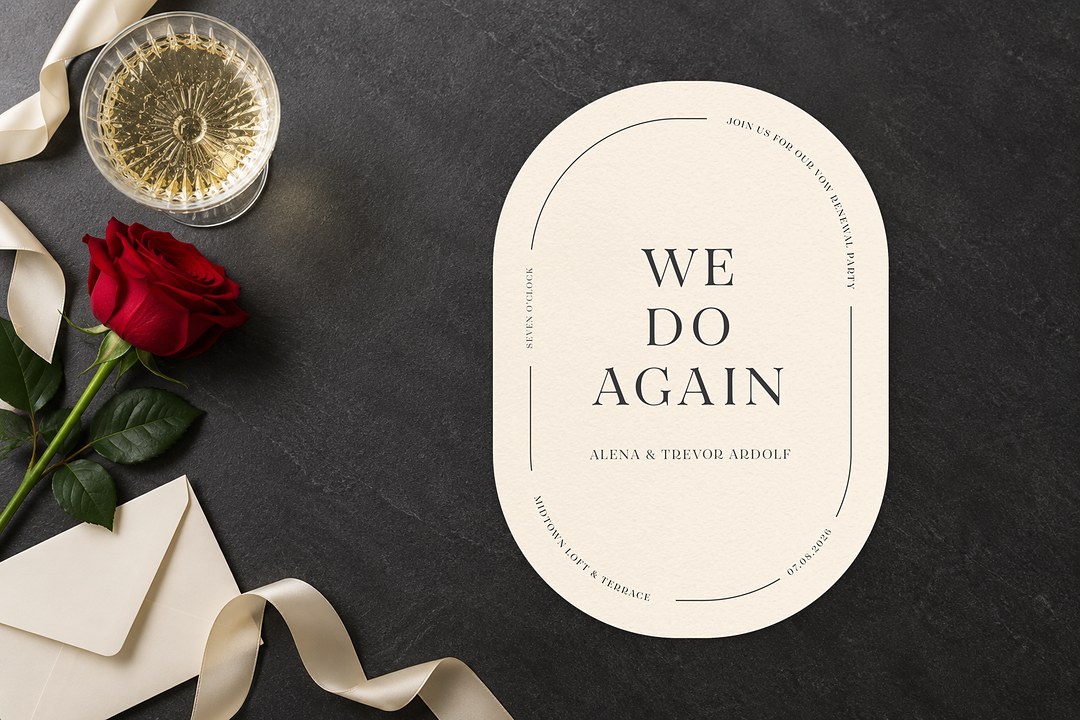 Paperlust All Minimal oval invitation with WE DO AGAIN sits on slate beside a red rose, cream envelope, ivory ribbon and champagne coupe.