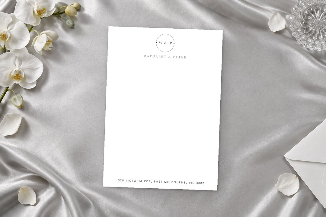 Paperlust Arch Lines notecard with an M & P monogram and Margaret & Peter lies on silver satin with orchids and an envelope.
