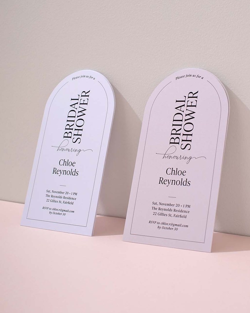 Two white arch shaped bridal shower invitations with black borders lean against a pale gray wall on a blush pink surface.