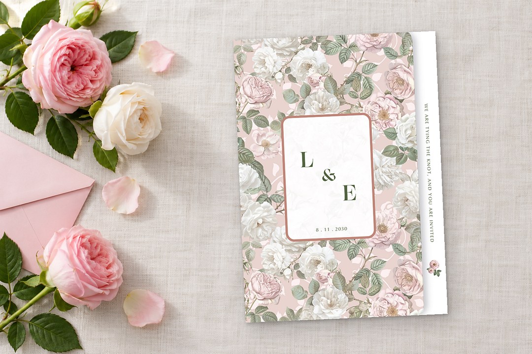 Paperlust Briar Rose folded invitation with pink and white roses lies on off white linen beside cream roses, pink envelope.