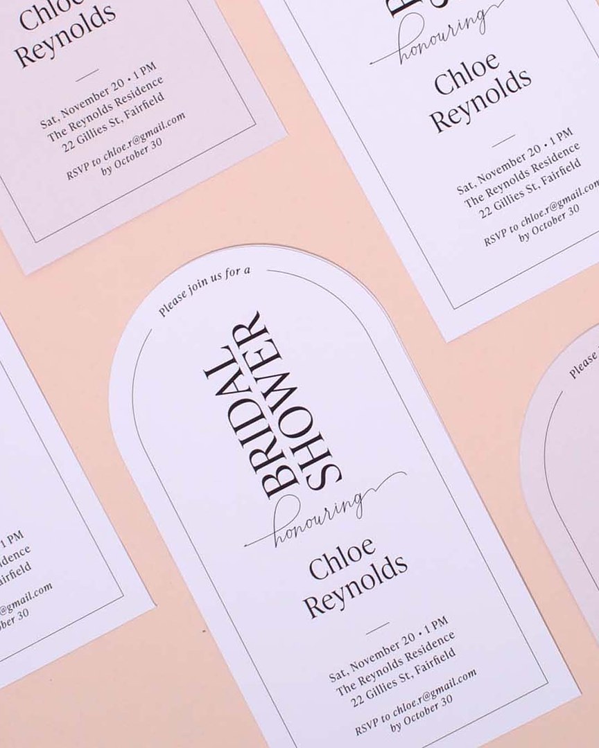 Arched white and blush bridal shower invitations with thin black border lines lie in rows on a peach surface.