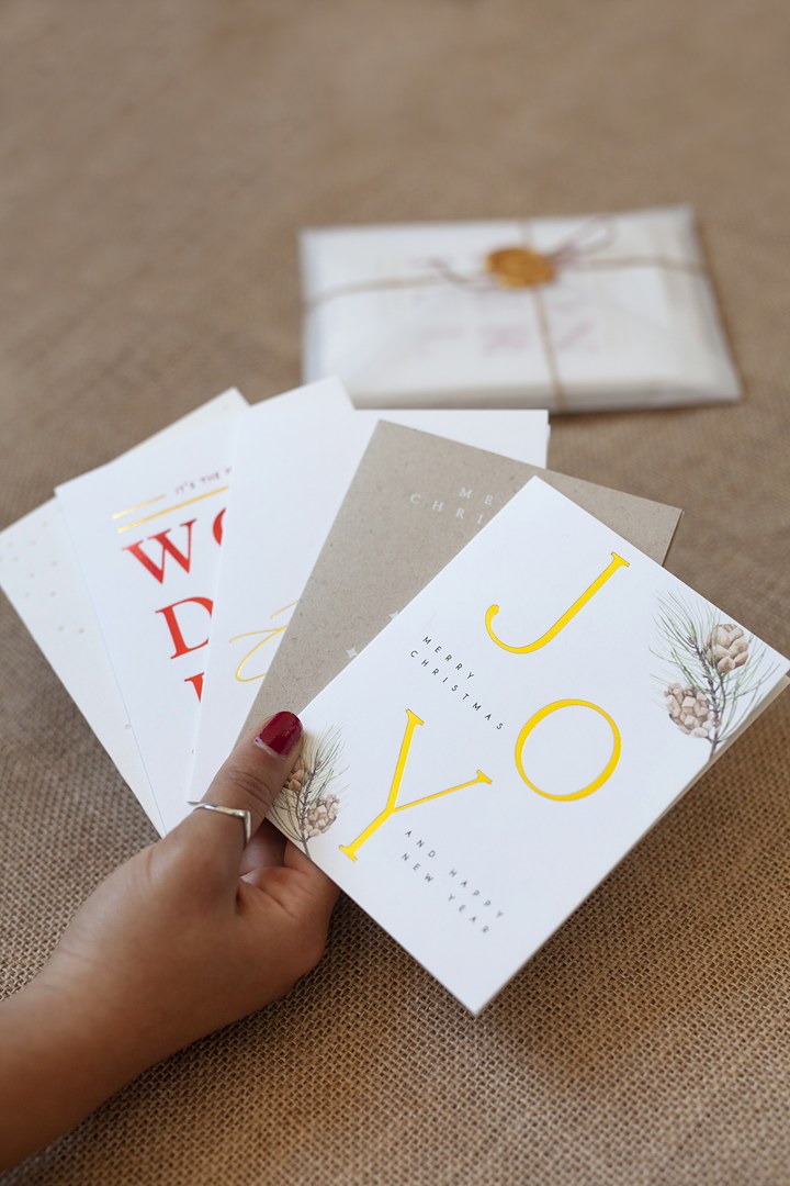 A hand with red nail polish and silver ring fans five Christmas cards on hessian cloth, with JOY in gold foil on the front card.