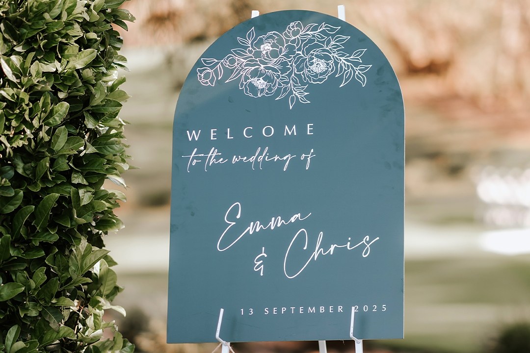 Teal arch shaped wedding sign with white lettering and a peony drawing stands on a white easel on a lawn beside a hedge.