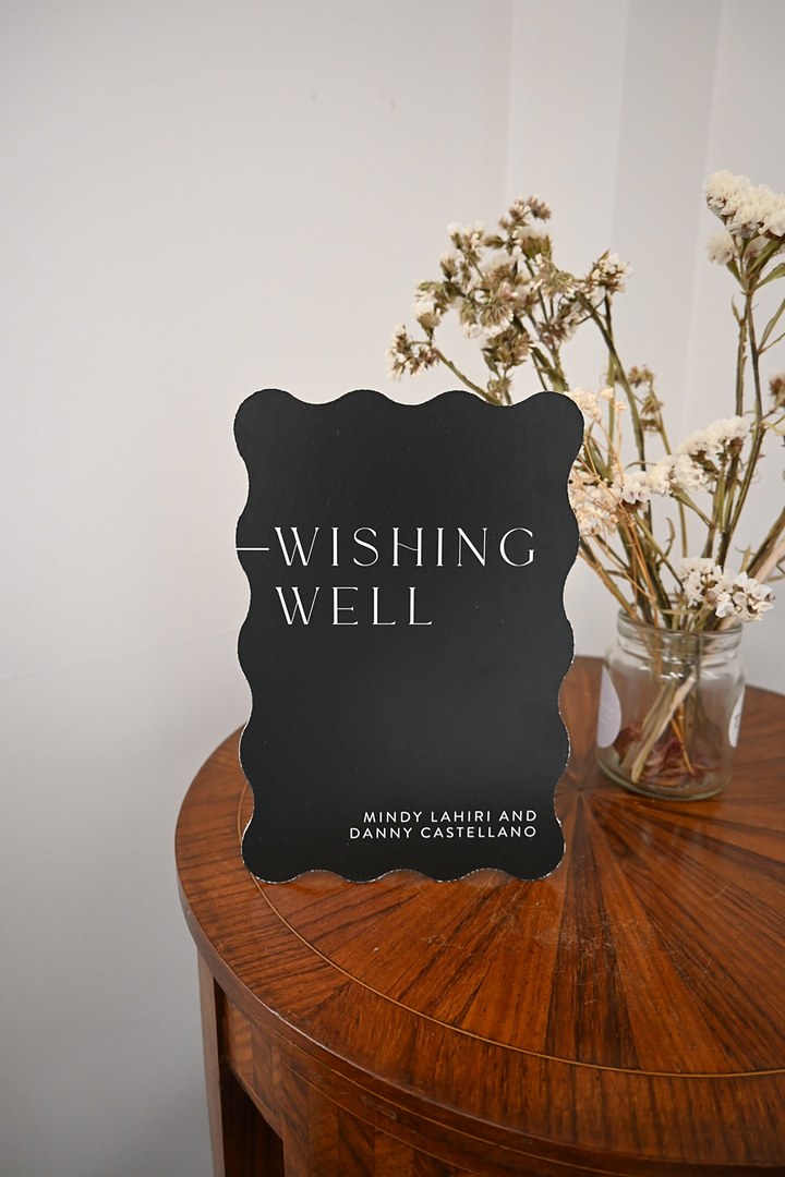 A black sign with a wavy die cut edge stands on a round wooden side table by a white wall, with a jar of dried pale flowers.