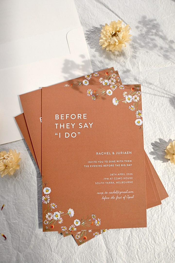 Paperlust Dancing Daisy terracotta invitations with white daisy corners lie on crinkled paper beside an envelope and flowers.