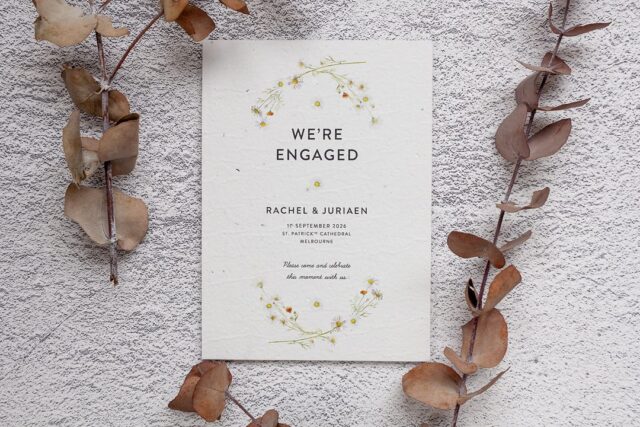 An engagement card with watercolor daisies and orange wildflowers sits on rough plaster beside dried eucalyptus stems.