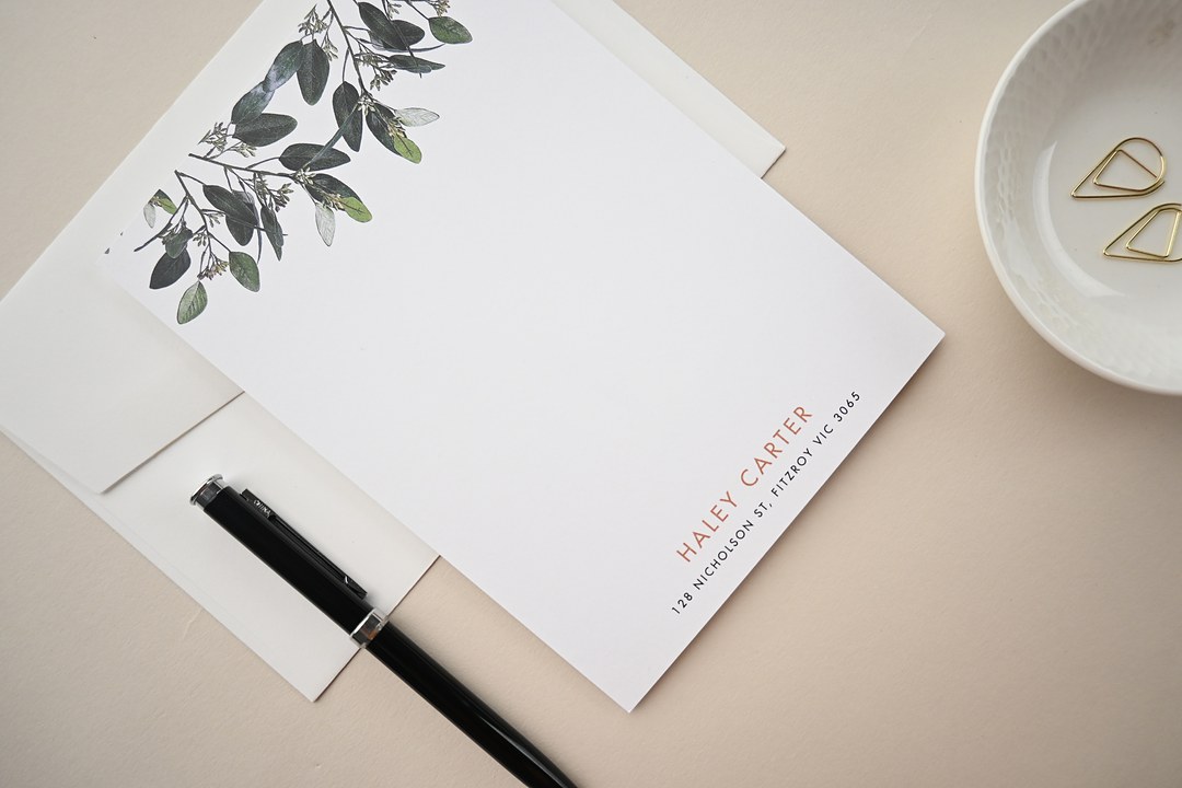 Paperlust Garden Estate notecard lies on an ivory envelope with eucalyptus leaves, a black pen and gold paper clips.