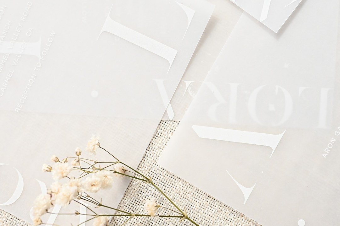 Paperlust Lissom overlapping vellum invitations in white ink lie on linen with dried cream flowers and white Lora letters.