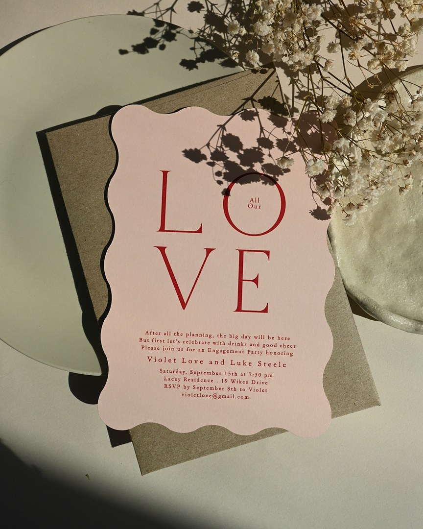 Paperlust Love Notes blush pink invitation with a wavy edge and red LOVE text lies on a kraft envelope and pale plate near dried flowers.