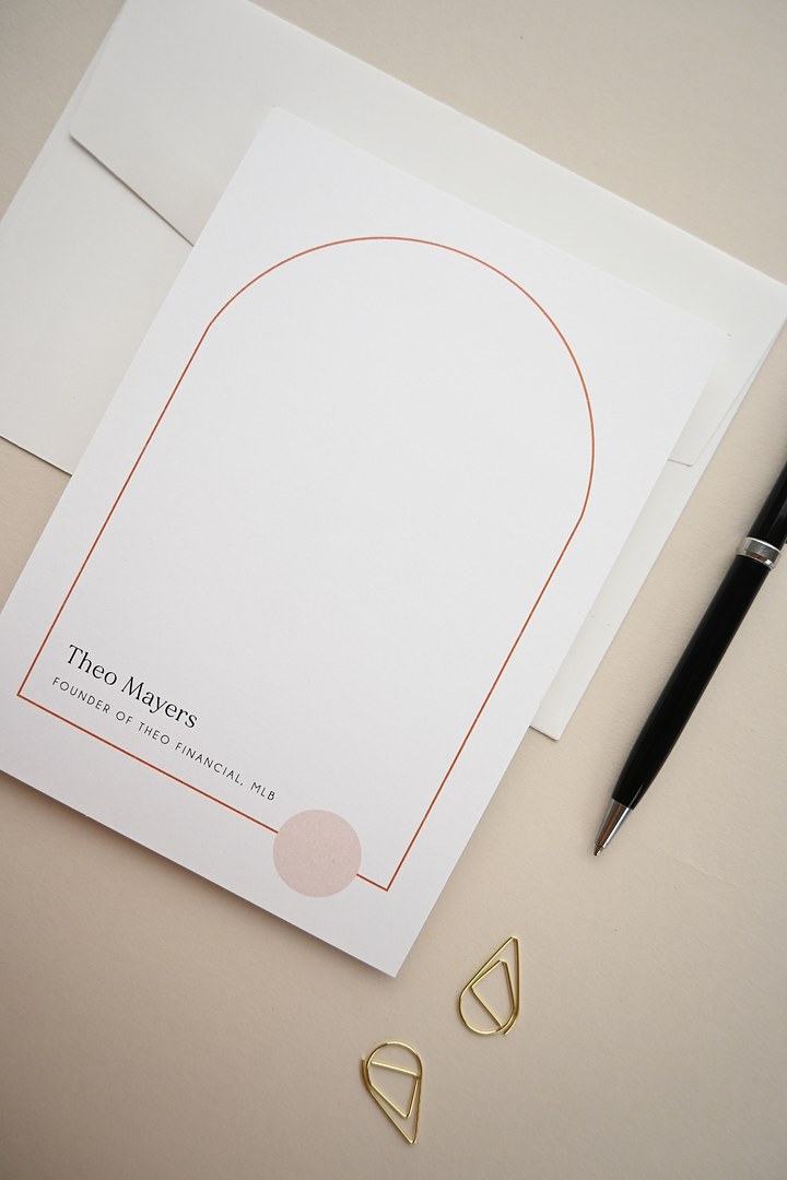 Paperlust Milos is a white notecard with a copper orange arch and pink circle on an envelope beside a pen and gold paper clips.