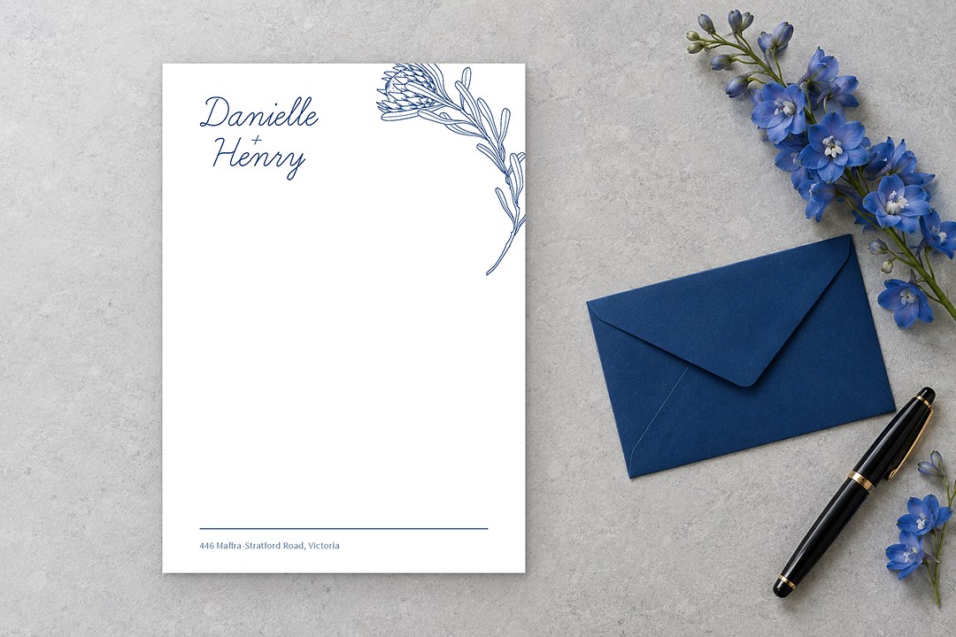 Paperlust Playful Proteas card with navy protea drawing lies on pale stone beside blue envelope and two delphinium stems.