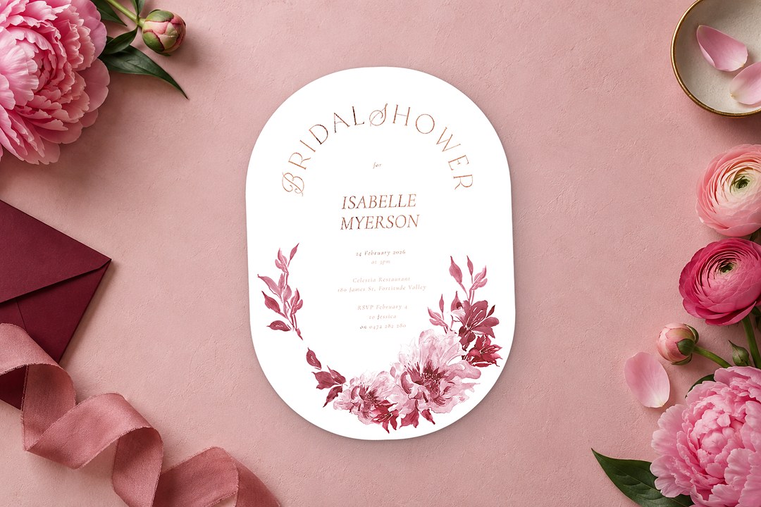 Paperlust Puspa white oval bridal shower invitation with rose gold lettering rests on pink beside peonies and a burgundy envelope.