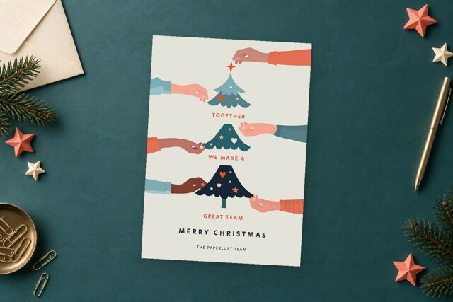 Paperlust Teamwork card on a deep teal desk is surrounded by a cream envelope, fir sprigs, coral and cream paper stars, and a brass pen.