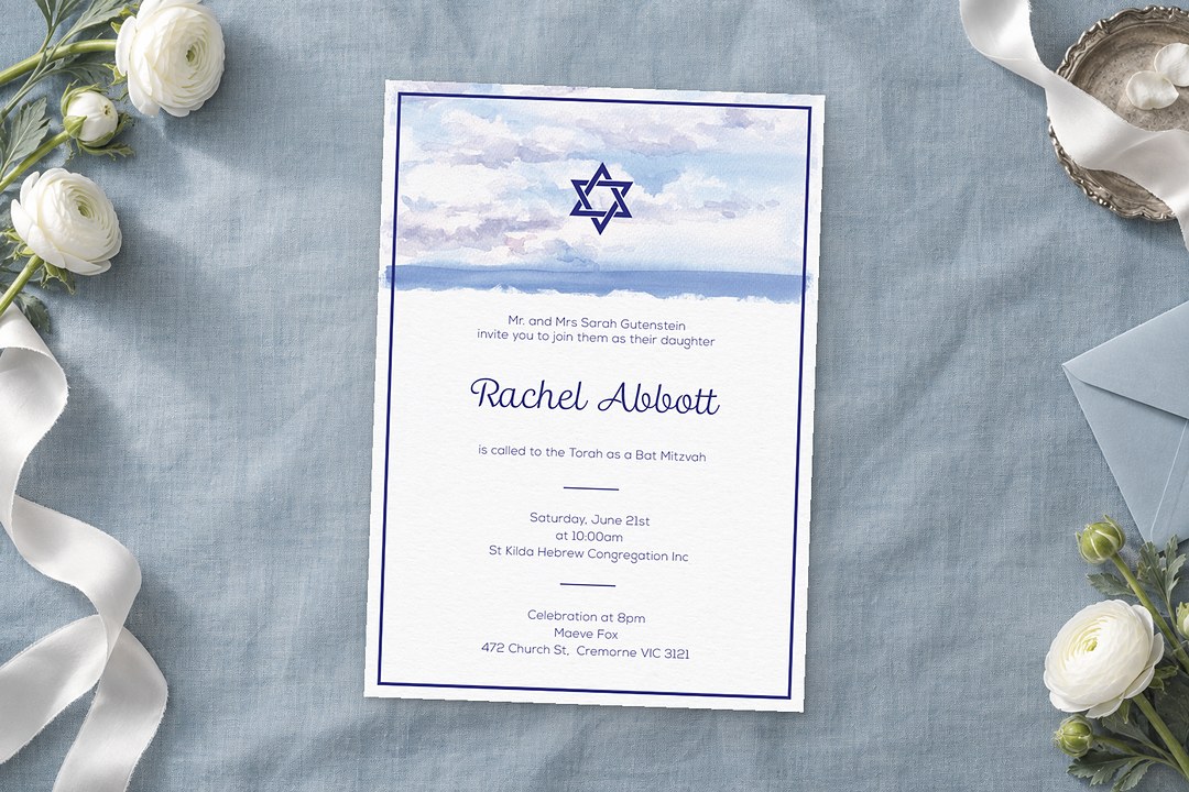 Paperlust The Day bat mitzvah invitation with a navy border lies on dusty blue linen beside white ranunculus and a pale blue envelope.