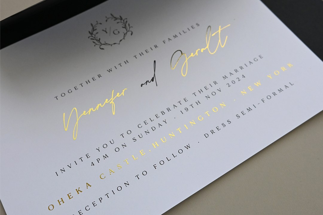 A white wedding invitation on a black envelope shows Yennefer and Geralt in gold foil with a Y G monogram in a leaf wreath.