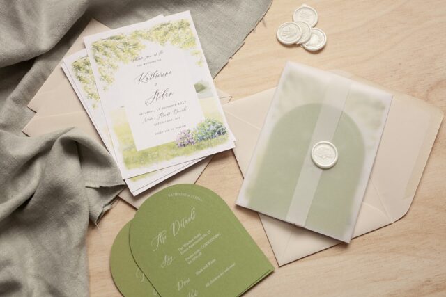 A stack of white invitations with watercolor branches and purple flowers rests on pale wood beside gray green linen.