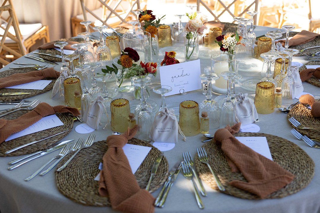 A round wedding reception table is set with woven placemats, terracotta napkins, amber glasses and a white card in a wire holder.