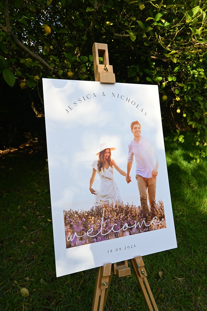 A white wedding welcome sign stands on a wooden easel by a lemon tree, showing a couple in white walking through lavender.