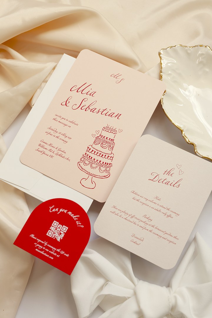 A blush pink wedding invitation with rounded corners lies on a white envelope over cream silk, printed with red script.