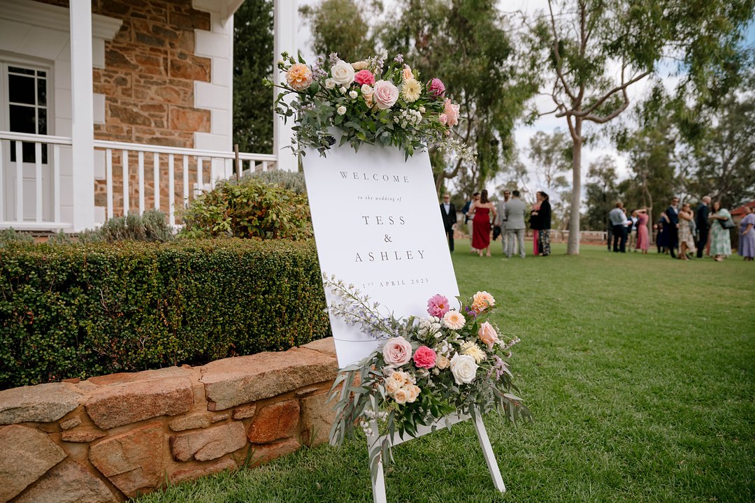 A white wedding sign on a white easel stands on a lawn beside a sandstone wall, decorated with pink, peach and cream roses.