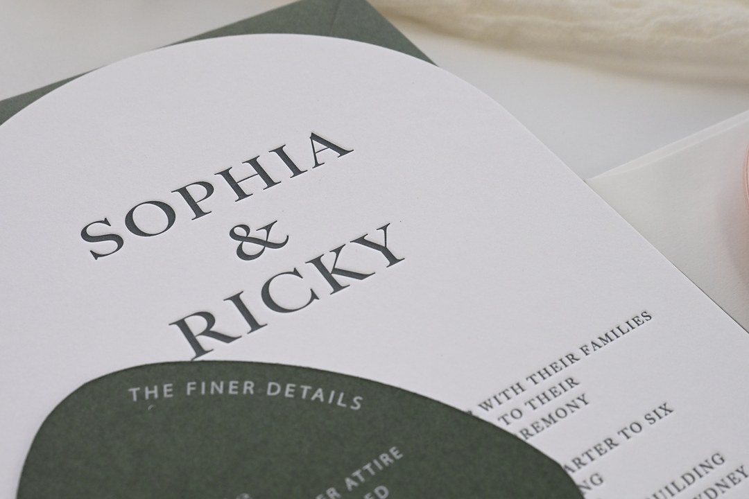 Thick white arch wedding invitation with Sophia & Ricky pressed in dark gray sits on a dark green envelope under a details card.