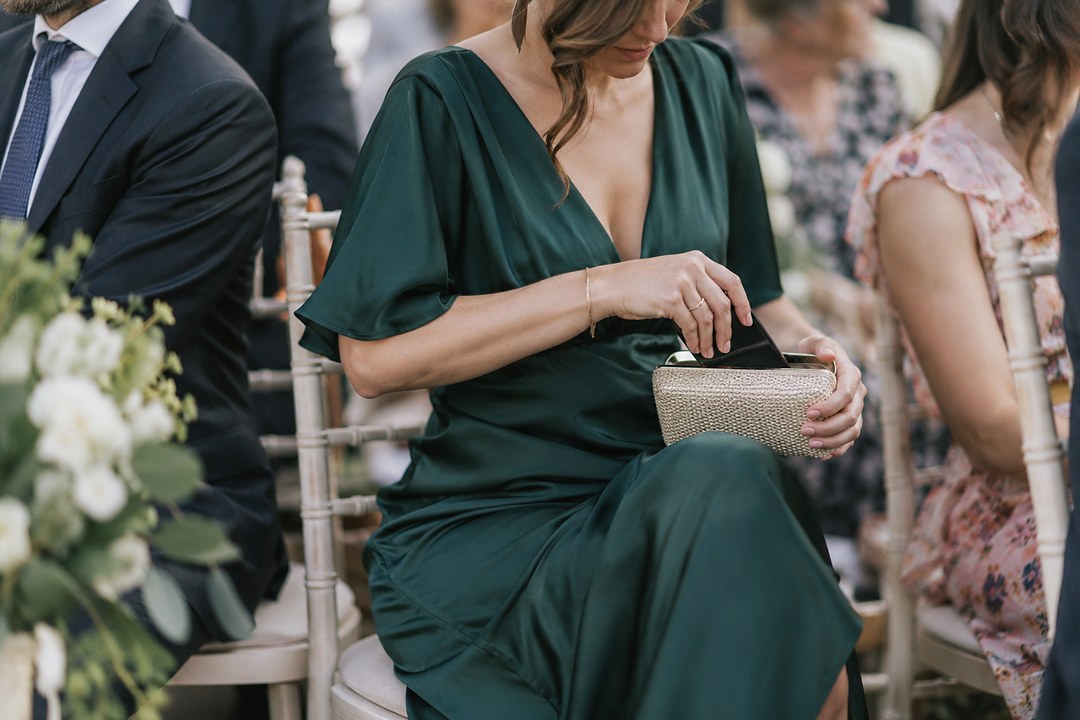 A seated guest in an emerald green satin dress slides a dark phone into a gold clutch on her lap among other ceremony guests.