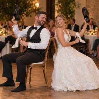 A bride and groom laugh back to back on wooden chairs on a dance floor, each holding a white heel and a black dress shoe.