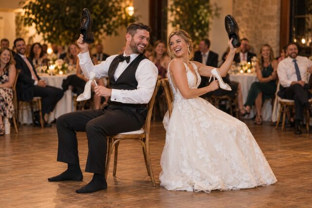 A bride and groom laugh back to back on wooden chairs on a dance floor, each holding a white heel and a black dress shoe.