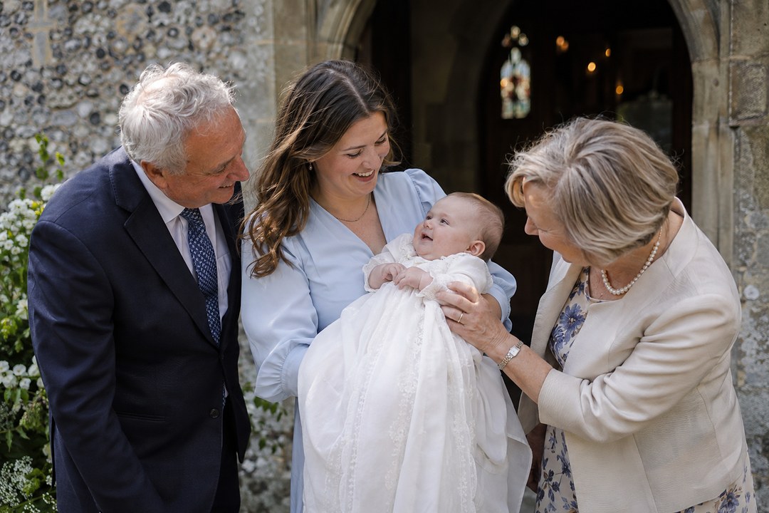 A young woman in a pale blue dress holds a smiling baby in a white gown outside a stone church while a man in navy smiles.