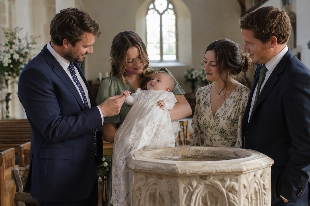 A woman in sage green cradles a baby in white lace beside a carved stone font while a man in navy holds the baby's hand.